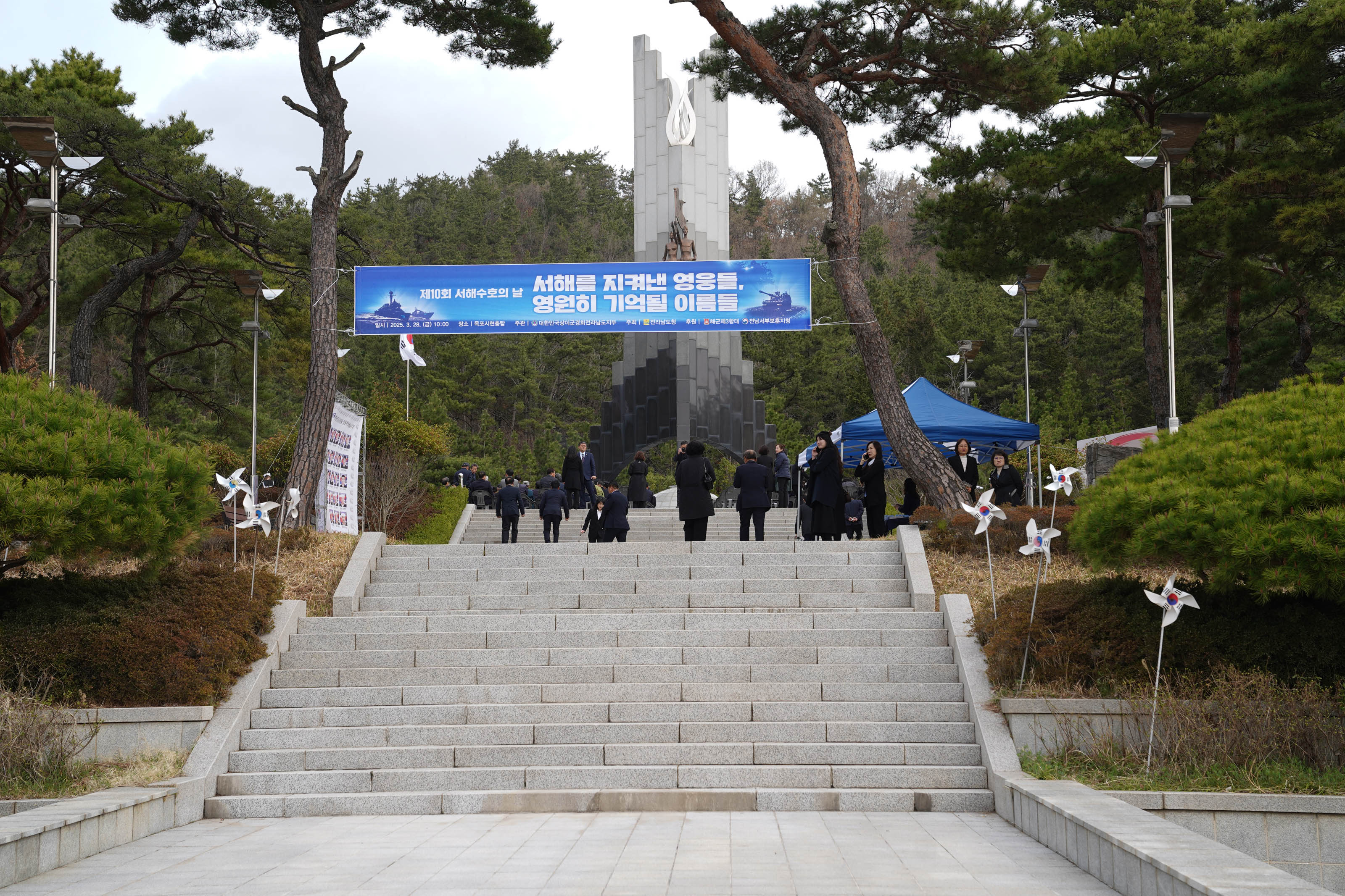 ‘제10회 서해수호의 날’ 기념식 현장. 기념탑을 향해 이어지는 계단 위에 현수막이 걸려 있으며, 현수막에는 “서해를 지켜낸 영웅들, 영원히 기억할 이름들”이라는 문구가 적혀 있다. 아래에는 행사 정보가 포함되어 있으며, “2025. 3. 28.(금) 10:00 / 전라남도·목포시 공동 주관 / 현충탑 앞”이라 쓰여 있다. 좌우로 태극기와 행사 참석자들이 보이고, 풍차 모양 장식에는 태극 문양이 붙어 있다.
