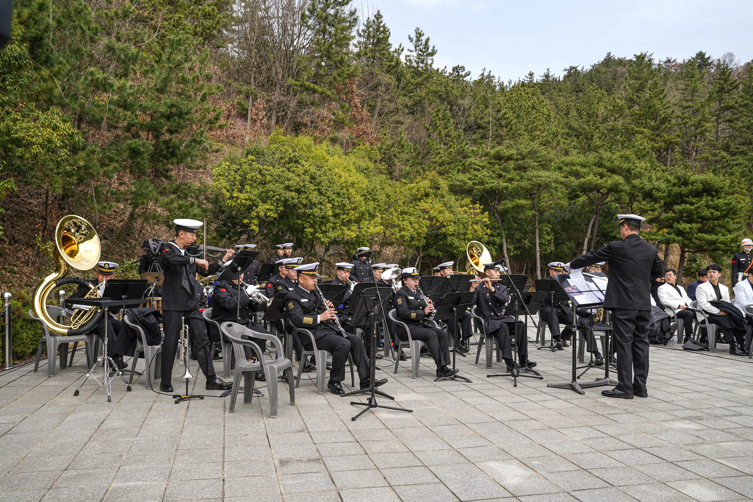 숲으로 둘러싸인 야외 공간에서 해군 군악대가 제10회 서해수호의 날 기념식을 위해 연주를 준비 중이다. 연주자들은 제복을 입고 각자 악기를 연주하거나 대기하고 있으며, 지휘자는 악보를 보며 지휘하고 있다. 악기에는 튜바, 클라리넷, 트럼펫 등이 보인다.