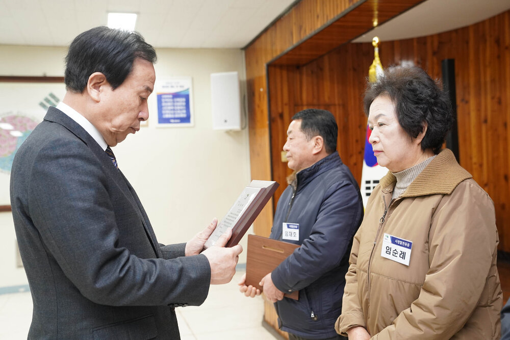 2월 정례조회에서 정장입은 남성이 어두운 베이지 자켓을 입은 여성에게 패를 건내고 있다.