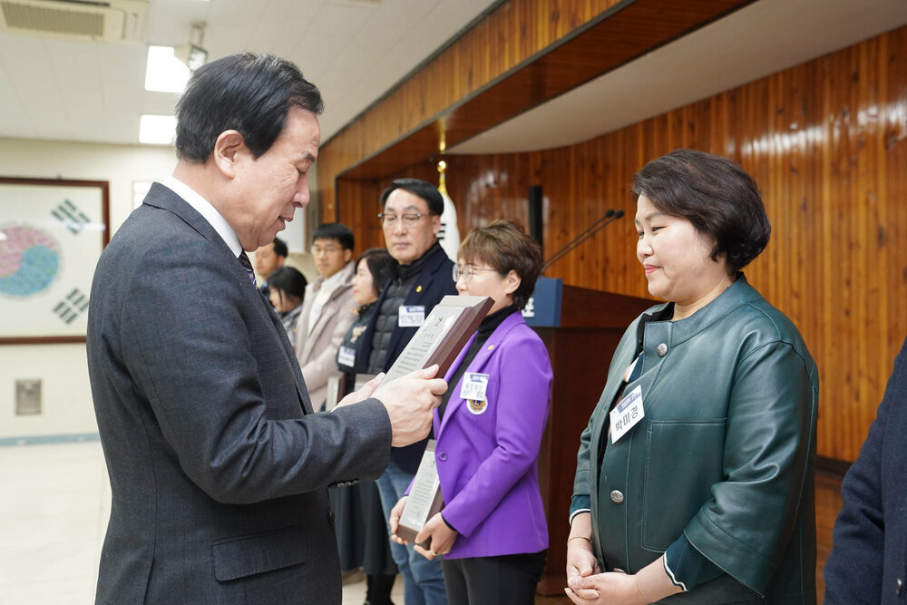 2월 정례조회에서 정장입은 남성이 어두운 초록빛의 가죽재킷을 입은 여성에게 패를 건내고 있다.
