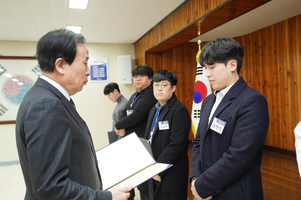 2월 정례조회에서 정장입은 남성이 펄 도트 무늬가 들어간 검정 자켓을 입은 남성에게 상장을 건내고 있다.