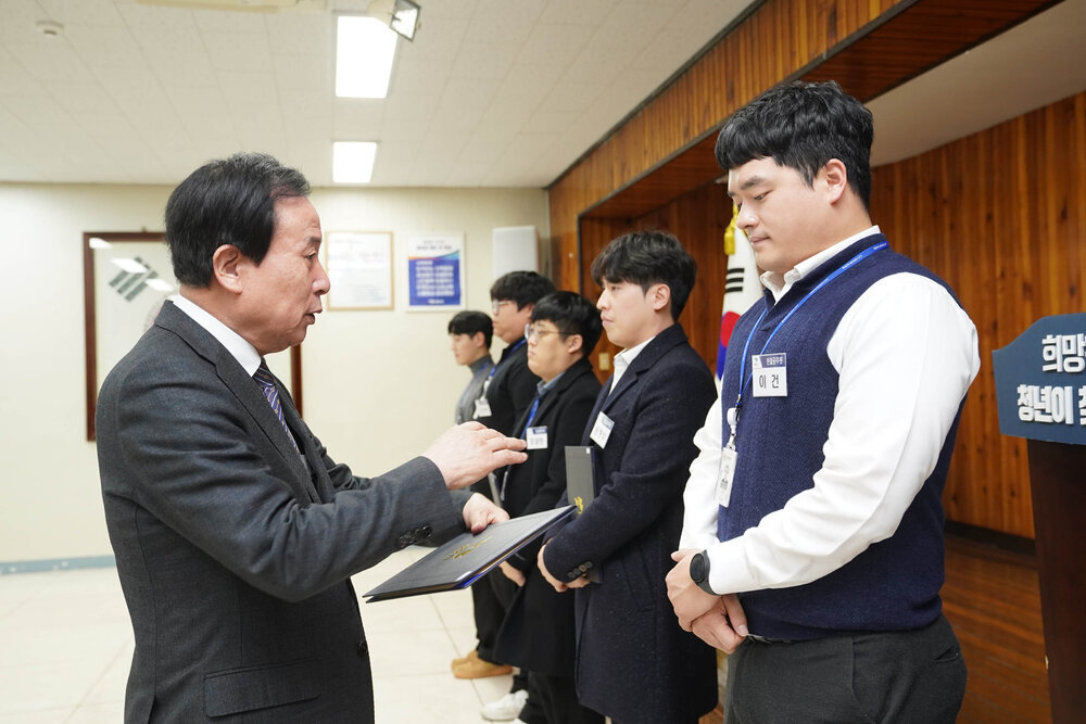 2월 정례조회에서 정장입은 남성이 흰 셔츠에 남색 베스트를 입은 남성에게 상장을 건내고 있다.