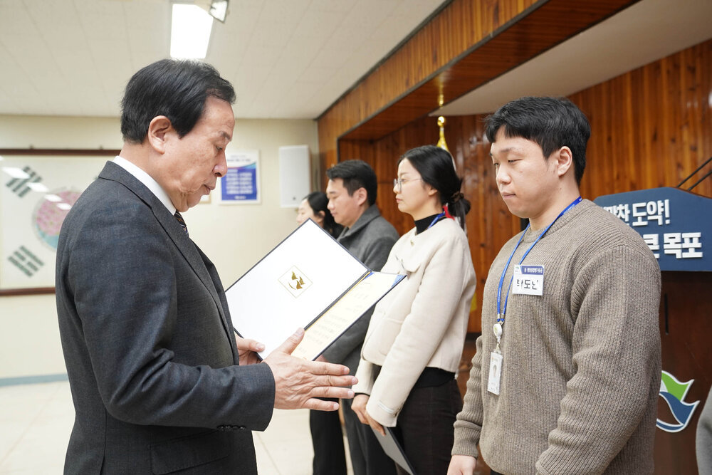 2월 정례조회에서 정장입은 남성이 갈색 니트를 입은 남성에게 상장을 건내고 있다.