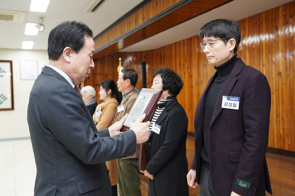 3월 정례조회에서 정장입은 남성이 어두운 보라색 자켓을 입은 남성에게 패를 건내고 있다.