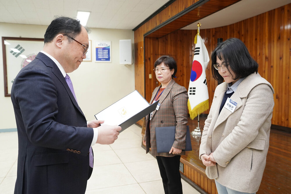 4월 정례조회에서 정장을 입은 남성이 베이지 코트를 입은 여성에게 표창장을 전달하는 모습