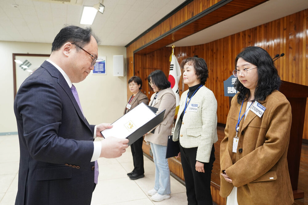 4월 정례조회에서 정장을 입은 남성이 갈색 재킷과 안경을 착용한 여성에게 표창장을 전달하는 모습