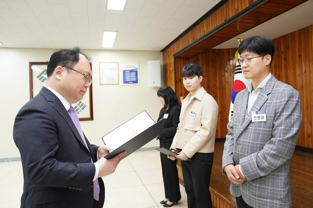 4월 정례조회에서 정장을 입은 남성이 체크무늬 재킷을 입은 안경 쓴 남성 직원에게 임용장을 전달하는 모습