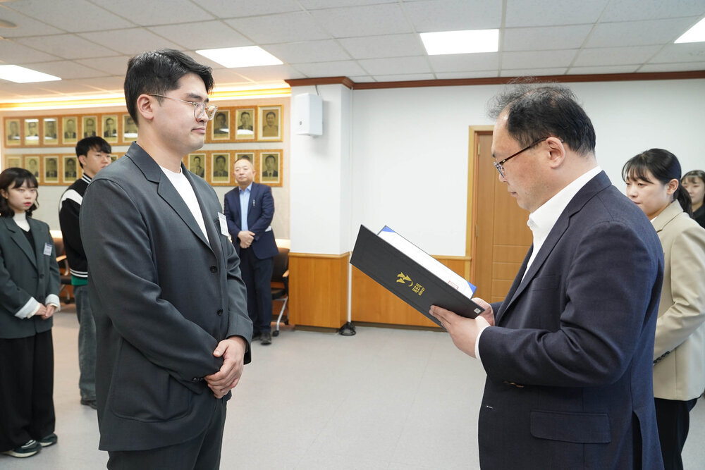 신규자 임용식에서 정장을 입은 남성이 검정 정장을 입은 안경 쓴 남성 직원에게 임용장을 전달하는 모습