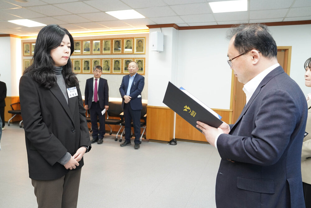 신규자 임용식에서 정장을 입은 남성이 검정 재킷과 회색 니트를 입은 여성 직원에게 임용장을 전달하는 모습