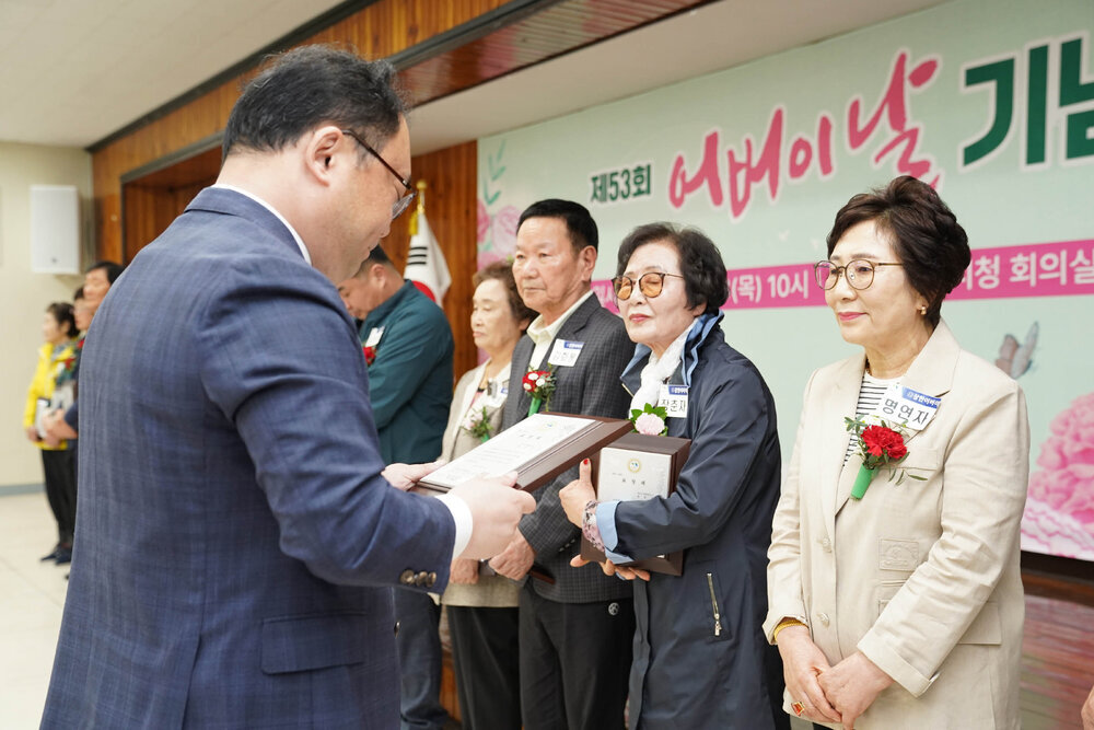 정장을 입고 안경을 낀 남성이 베이지색 자켓을 입은 여성에게 패를 수여하고 있다