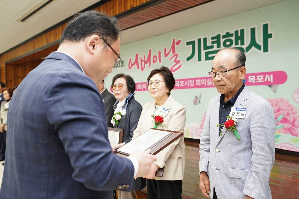 정장을 입고 안경을 낀 남성이 연보라색 자켓을 입은 남성에게 패를 수여하고 있다