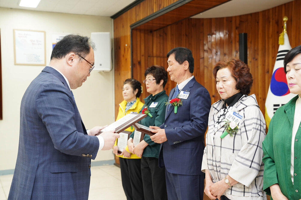 정장을 입고 안경을 낀 남성이 검은색 줄무늬 체크 패턴이 들어간 크림색 셔츠를 입은 강덕순에게 패를 수여하고 있다