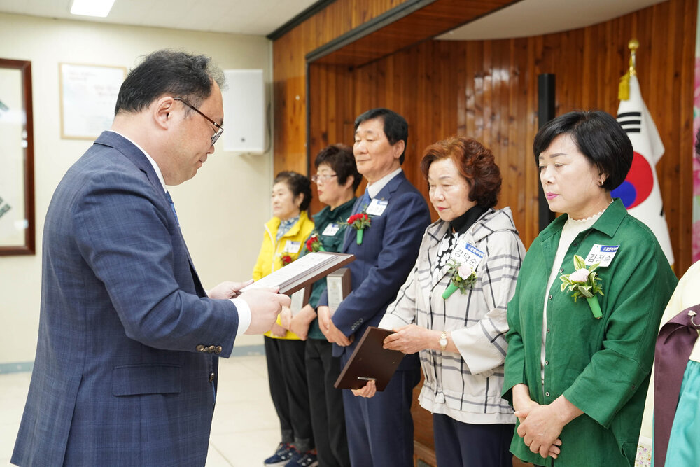 정장을 입고 안경을 낀 남성이 초록색 셔츠를 입은 여성에게 패를 수여하고 있다