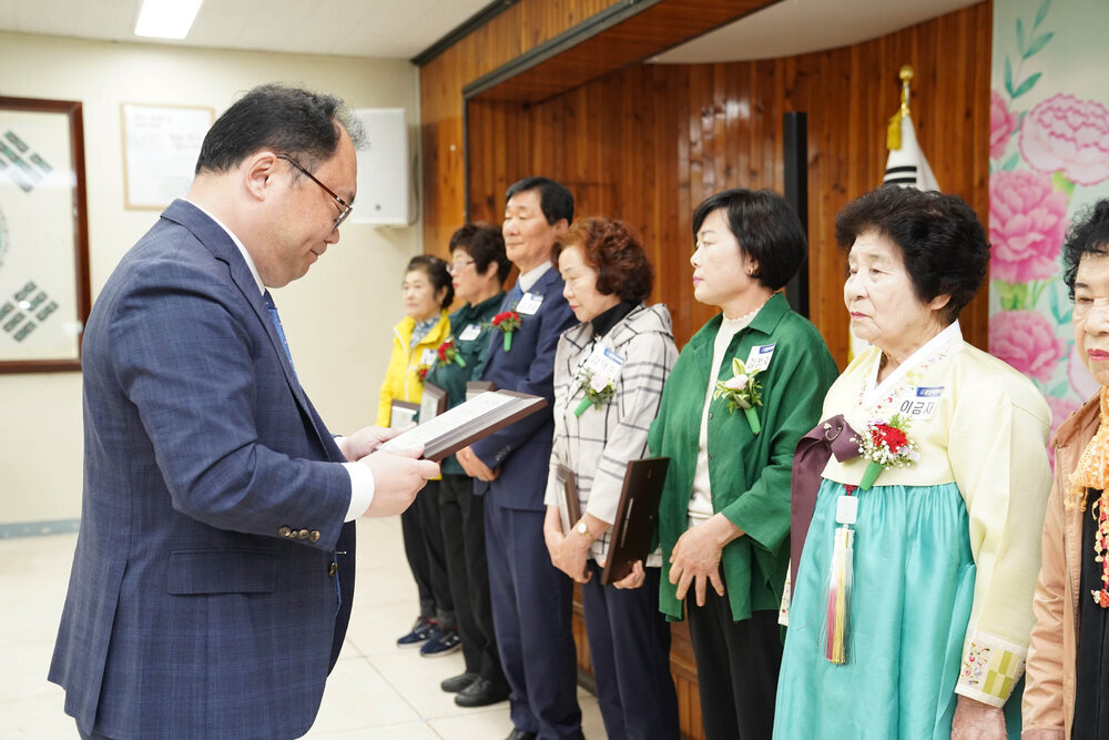 정장을 입고 안경을 낀 남성이 연노랑 저고리에 민트색 한복 치마를 입은 이금자에게 패를 수여하고 있다