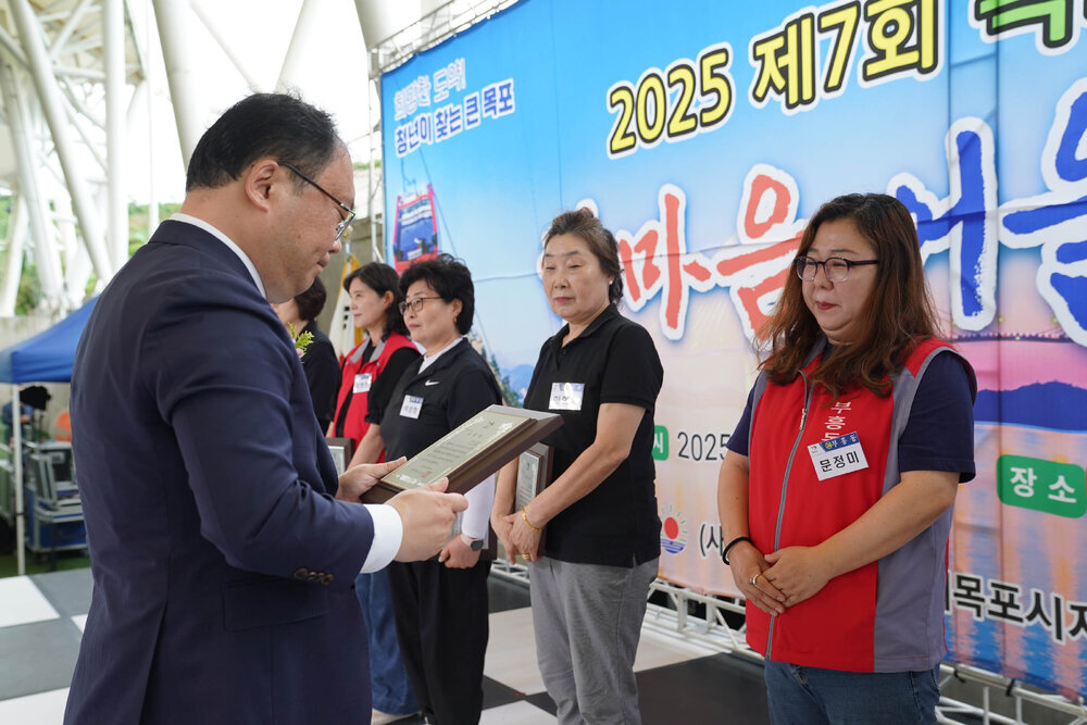 정장을 입고 안경을 낀 남성이 곤색 반팔티에 빨간색 작업조끼를 입은 문정미에게 패를 수여하고 있다