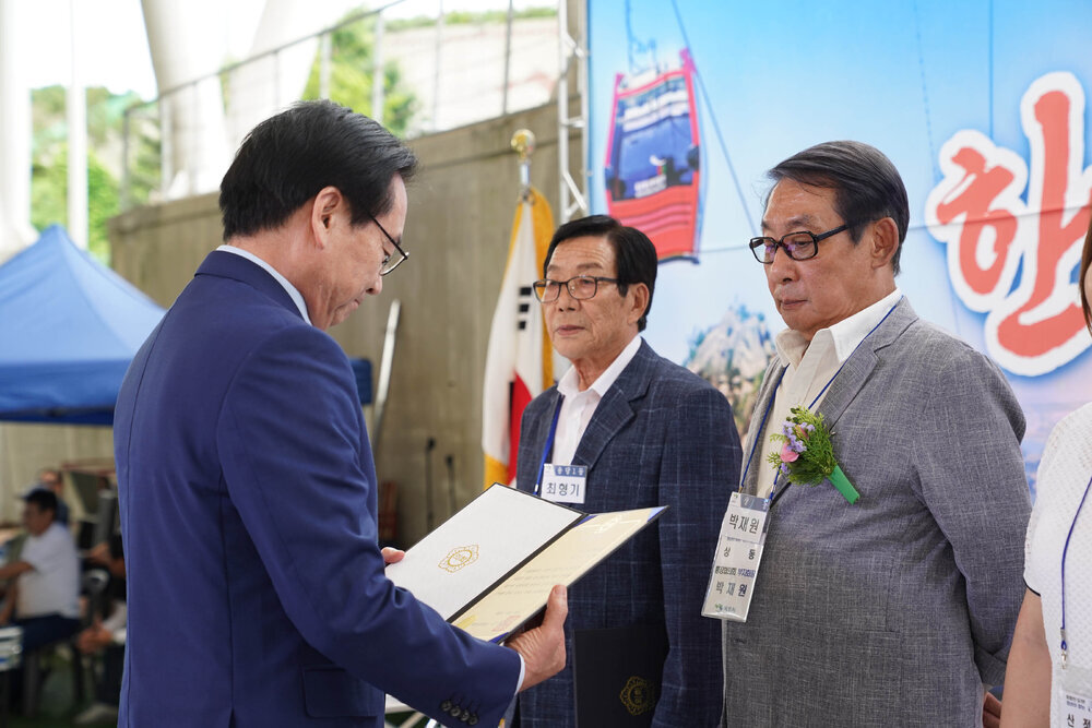곤색 정장을 입고 안경을 낀 남성이 회색 자켓을 입고 카라에 꽃을 단 박재원에게 상장을 수여하고 있다