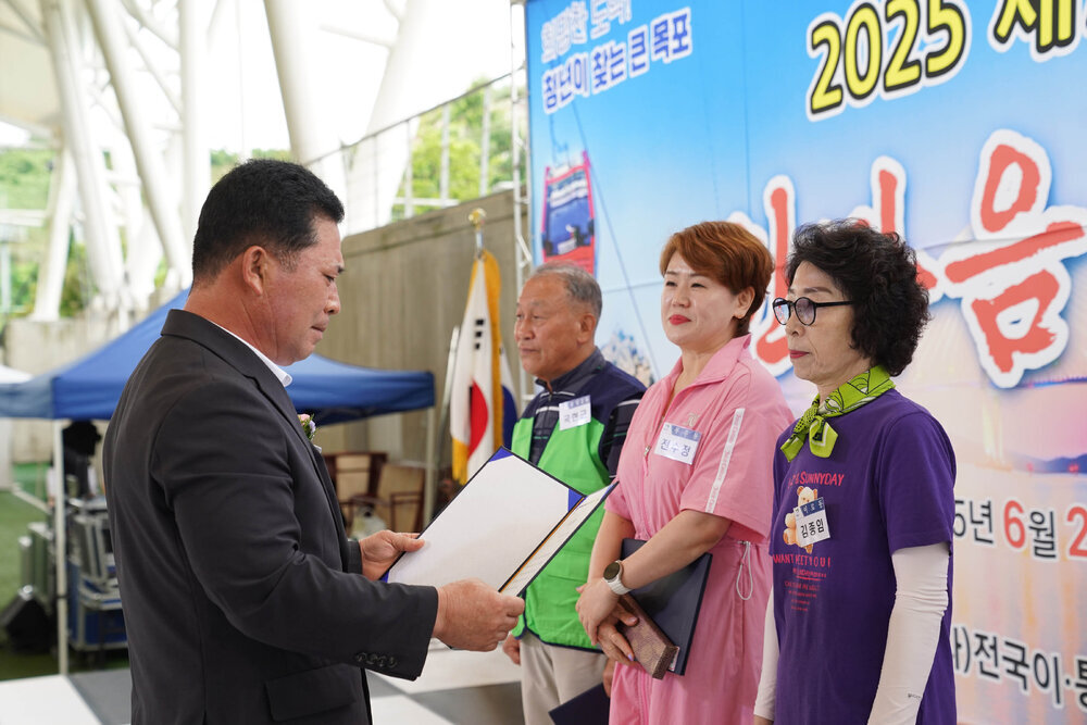 곤색 정장을 입고 안경을 낀 남성이 흰색 팔토사를 끼고 보라색 반팔티를 입고 연두색 스카프를 두른 여성에게 상장을 수여하고 있다
