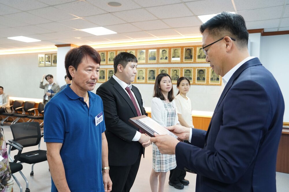 정장 차림의 관계자가 파란색 반팔 티셔츠를 입은 남성 수상자에게 표창패를 전달하며 읽고 있는 모습이다.