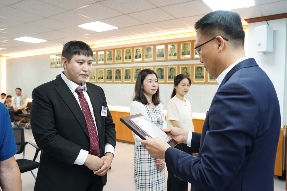 정장 차림의 관계자가 검은 정장을 입은 남성 수상자에게 표창패를 전달하는 모습이며 뒤에 다른 수상자들이 대기하고 있다.