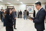 2026년 상반기 4급이하 정기인사로 정장을 입은 남성이 실내 행사장에서 한 여성에게 상장을 전달하며 내용을 읽고 있고, 여성은 파마머리에 검은색 정장 자켓을 입고 사원증을 착용한 채 두 손을 모은 상태로 서 있으며 뒤쪽으로 참석자들이 줄지어 서서 행사를 지켜보고 있다.