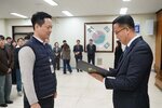 2026년 상반기 4급이하 정기인사로 정장을 입은 남성이 실내 행사장에서 한 남성에게 상장을 전달하며 내용을 읽고 있고, 남성은 짧은 머리에 남색 경량 패딩 조끼를 입고 사원증을 착용한 채 두 손을 내린 상태로 서 있으며 뒤쪽으로 참석자들이 줄지어 서서 행사를 지켜보고 있다.