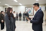 2026년 상반기 4급이하 정기인사로 정장을 입고 안경을 쓴 남성이 실내 행사장에서 한 여성에게 상장을 전달하며 내용을 읽고 있고, 여성은 회색 코트를 입고 서 있으며 뒤쪽에는 참석자들이 줄지어 서서 행사를 지켜보고 있다.